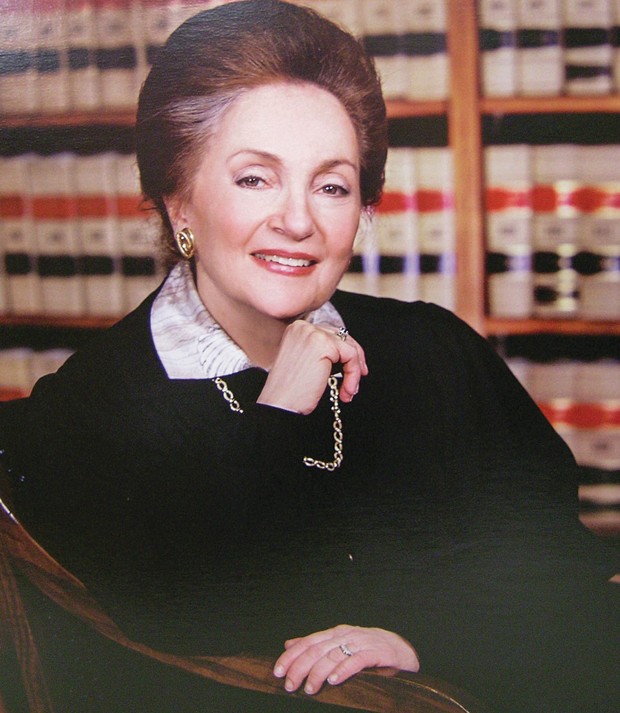 Portrait headshot photograph of The Honorable Dolores K. Sloviter L’56 smiling while she poses with her right hand underneath her chin area sitting on a wooden style chair wearing her black color judge gown, golden color earrings, golden color necklace, and silver color rings on both of her hands