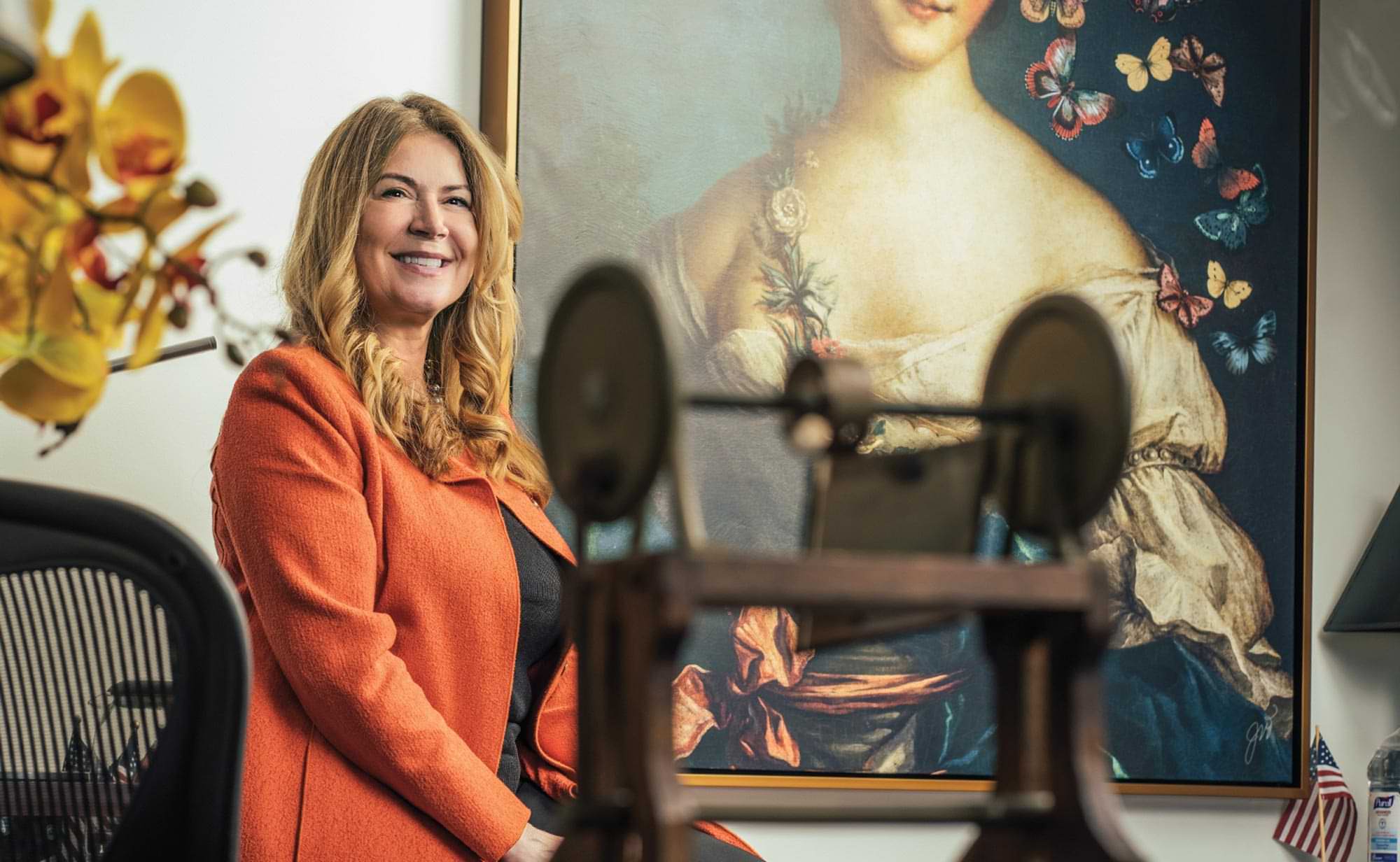 Kathi Vidal smiles while sitting at the edge of a table near a large hanging classical painting