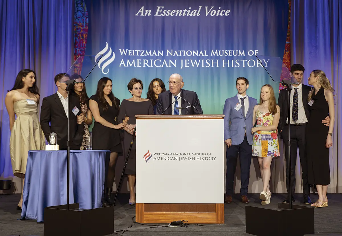 Portrait close-up indoor photograph view of Steve Cozen C’61, L’64 receiving the inaugural Only in America Humanitarian Award from the Weitzman National Museum of American Jewish History, the preeminent national museum dedicated to sharing Jewish stories that shaped the United States as he is speaking at a podium while behind him there are a group of people all posing and standing listening to him talk as he address an audience