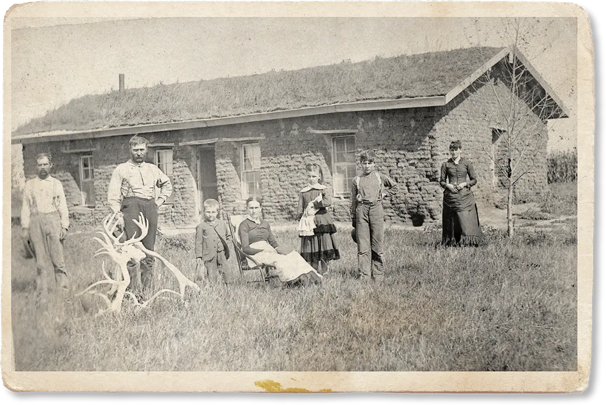 A sepia-toned photo shows six people in front of a stone building with a grassy roof.