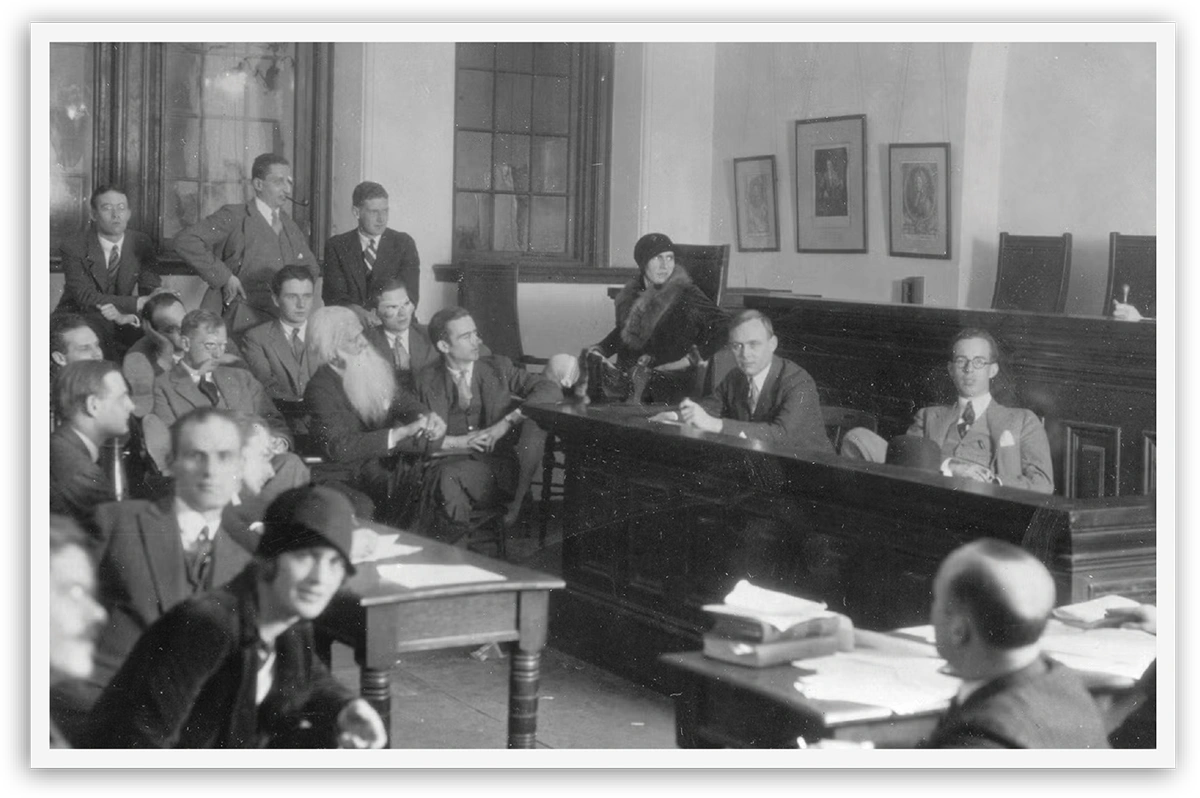 Black-and-white courtroom scene with people seated and standing, observing proceedings.